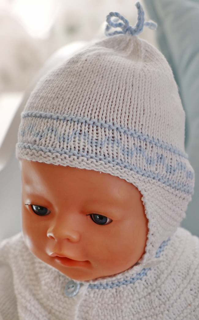 Knit a complementary hat in white with blue patterns to match the jacket. It includes functional earflaps and a button closure under the chin, secured with a knitted chord. A blue chord bow on the top adds a final touch of cuteness.
